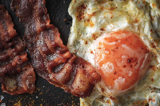 From above of sunny side up egg with fried bacon slices and condiments on dark tray — Stock Photo