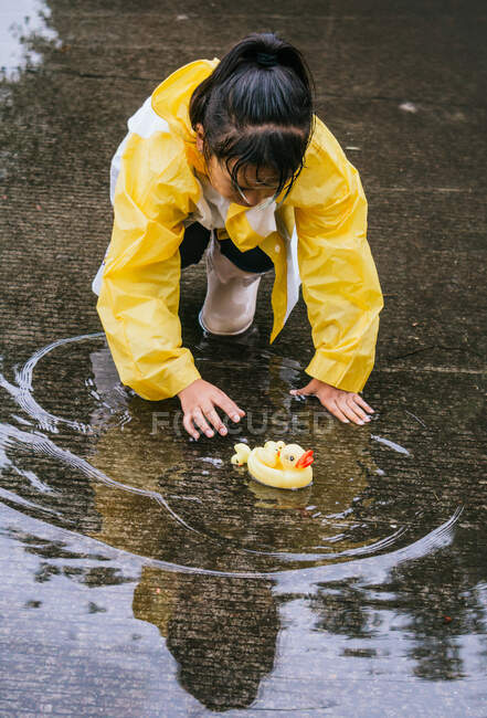 Criança étnica encantadora em capa de chuva brincando com patos de plástico refletindo em poça ondulada em tempo chuvoso — Fotografia de Stock