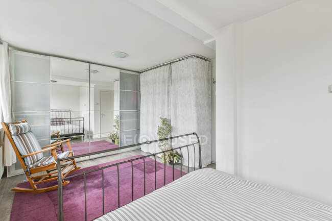 Cama com travesseiros no quarto claro espaçoso com poltrona colocada no tapete rosa perto da janela e no grande guarda-roupa espelhado — Fotografia de Stock