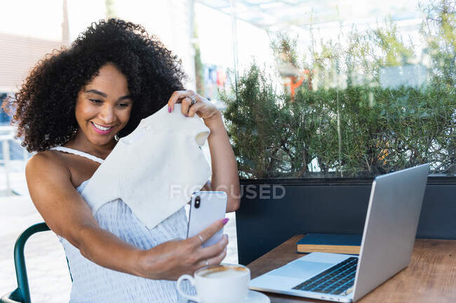 Positive pregnant African American female sitting at table with laptop and latte and taking self portrait with baby clothes — Stock Photo