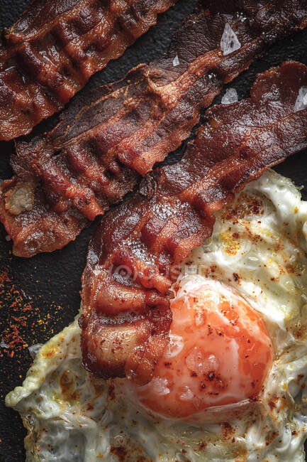 De cima do lado ensolarado acima do ovo com fatias de bacon fritas e condimentos na bandeja escura — Fotografia de Stock