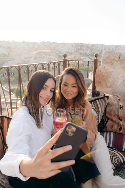 Alto ángulo de amigos étnicos felices sentados en almohadas mientras toman autorretrato en el teléfono celular cerca de la mesa con cámara de fotos instantánea y cóctel en la terraza en Capadocia, Turquía - foto de stock