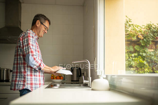 Seitenansicht des ruhigen glücklichen reifen Mannes, der schmutzige Teller wäscht, während er in der Küche neben der Spüle steht und Hausarbeit erledigt — Stockfoto