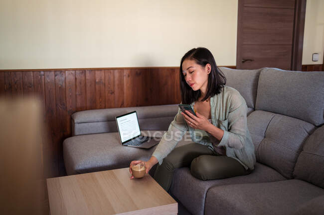 Young ethnic female with glass of coffee surfing internet on cellphone sitting in couch at home room near laptop — Stock Photo