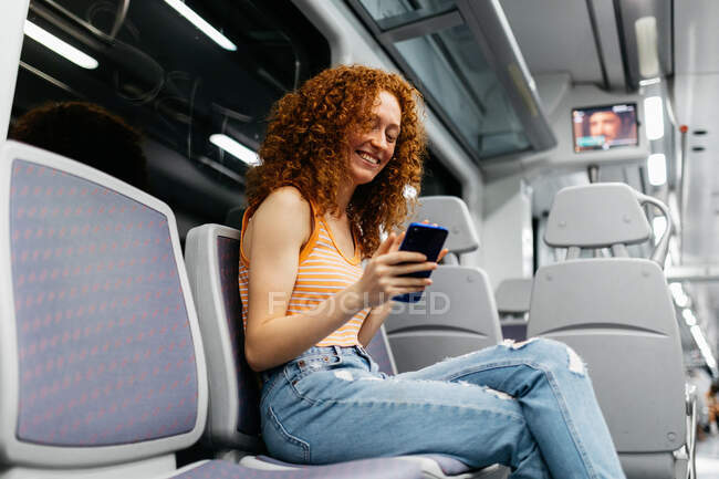 Mujer interesada con el pelo rizado en vaqueros rasgados mensajes de texto en el teléfono celular durante el viaje en tren durante el día - foto de stock