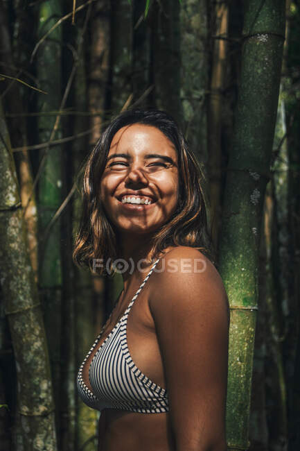 Side view of content young female traveler in swimwear with shadow on face looking away against bamboo twigs — Stock Photo