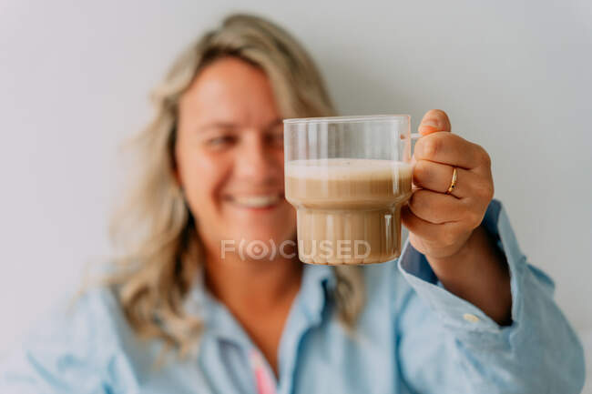 Conteúdo adulto loiro fêmea segurando xícara de café delicioso com espuma de leite no topo no fundo claro — Fotografia de Stock