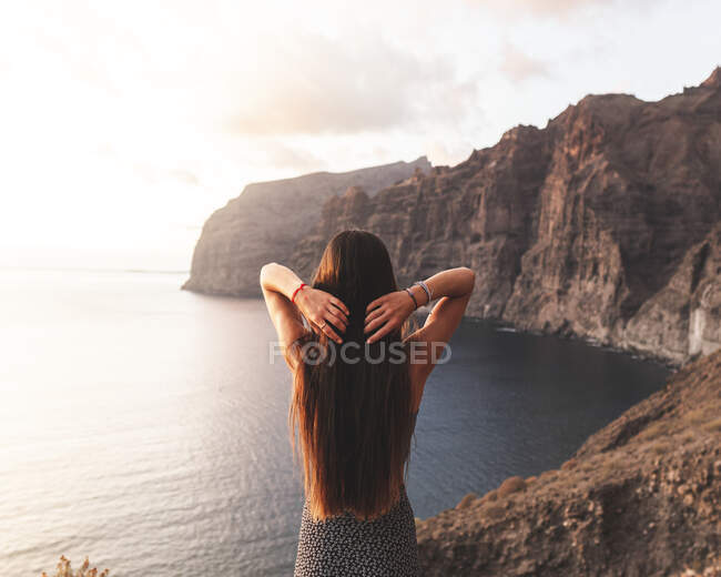 Vista posterior de una mujer anónima con el pelo largo y las manos detrás de la cabeza en la cresta contra el océano al atardecer en Tenerife España - foto de stock