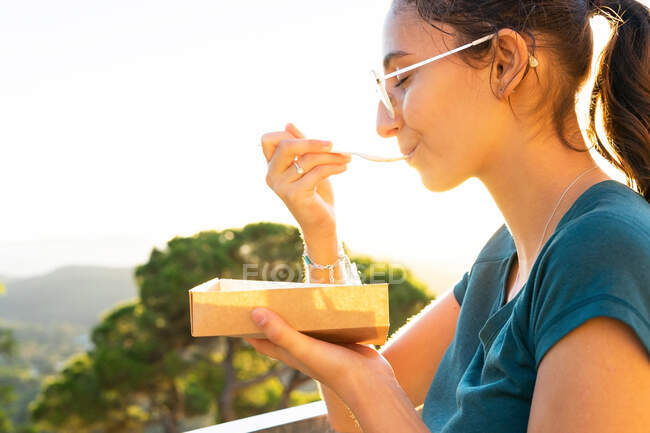 Side view of crop young female eating tasty Belgian waffles with whipped cream in takeaway box against mounts in back lit — Stock Photo