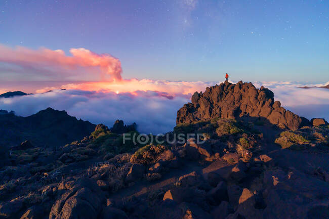 Nächtliche Landschaft mit einem ausbrechenden Vulkan im Hintergrund und einem Wolkenmeer, das die Berge in einer sternenklaren Nacht von einem bewachsenen und felsigen Berg und einer Person, die oben auf dem Kamm steht, bedeckt. Cumbre Vieja Vulkanausbruch auf La Palma Kanarische Inseln I — Stockfoto