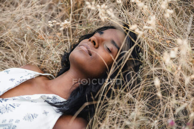 Achtsame erwachsene ethnische Hündin mit geschlossenen Augen und dunklen Haaren, die tagsüber auf trockenem Gras liegt — Stockfoto