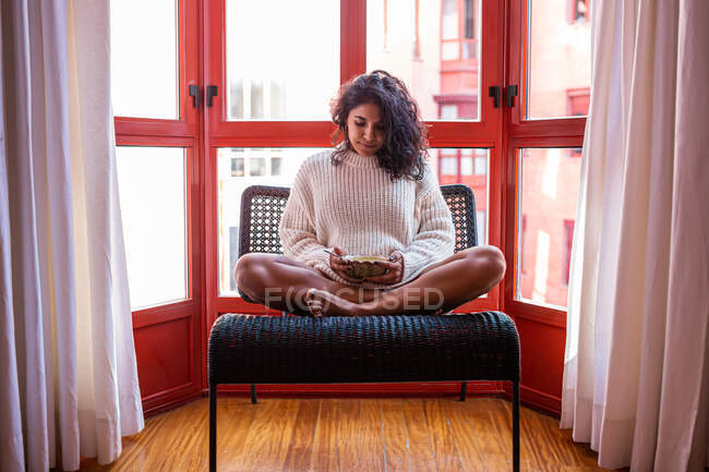 Full body of barefoot latin female sitting with crossed legs on chair and eating soup from bowl — Stock Photo