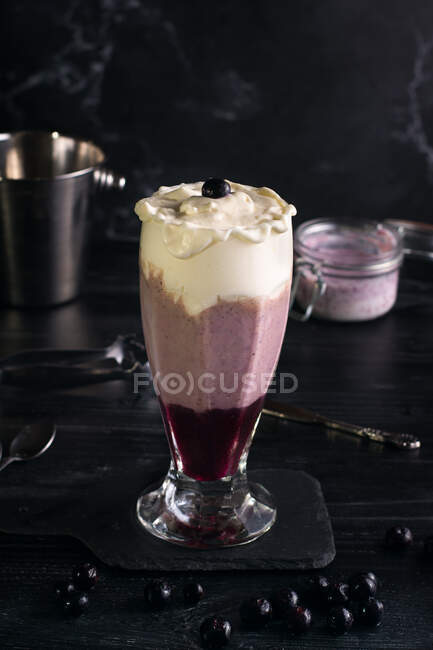 Glass of tasty smooth banana and blueberry drink with whipped cream on top on cutting board on dark background — Stock Photo