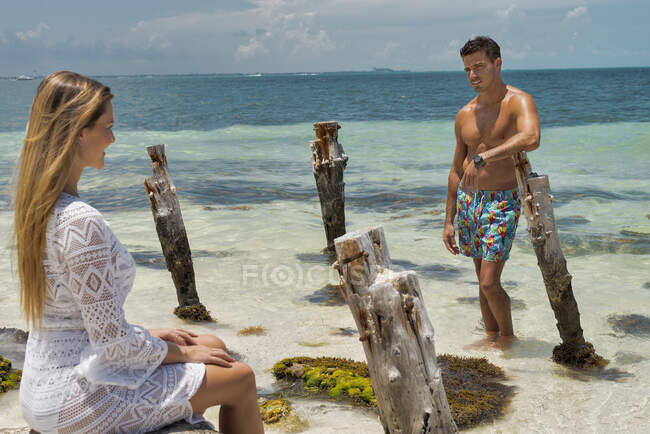Bonito cara com torso muscular bronzeado conversando com uma jovem encantadora em roupas de praia enquanto relaxam juntos durante a lua de mel romântica na costa tropical — Fotografia de Stock