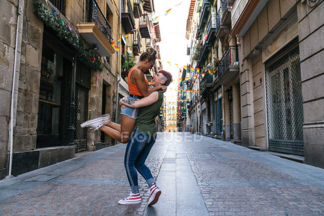 Vista lateral da moda jovem mulher tatuada carregando namorada homossexual enquanto rindo e olhando um para o outro na passarela urbana — Fotografia de Stock