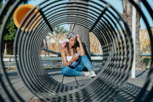 Cool ethnic female listening to song from headset while sitting with crossed legs against tunnel shaped construction — Stock Photo