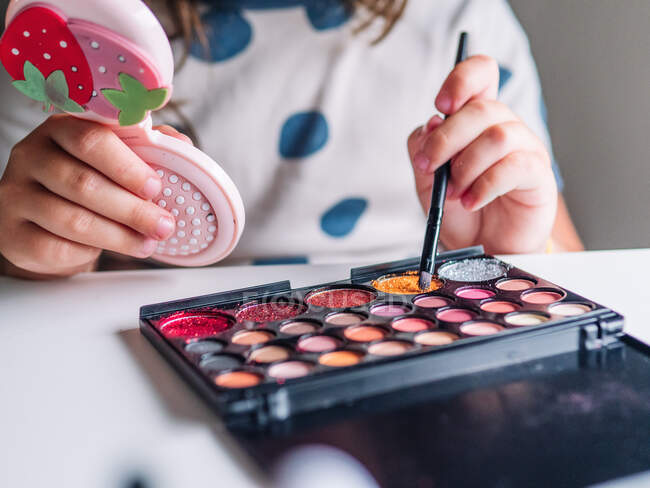 Cortar criança irreconhecível com aplicador e espelho tornando-se rosto à mesa com produtos cosméticos variados em casa — Fotografia de Stock