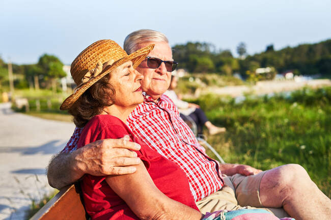 Vue latérale du couple âgé assis regardant loin sur un banc en bois et profitant de la journée d'été sur le rivage de l'étang — Photo de stock