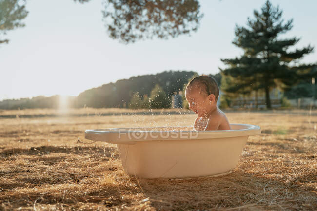 Side view of happy toddler child with toy sitting in plastic bath while playing with water in countryside — Stock Photo