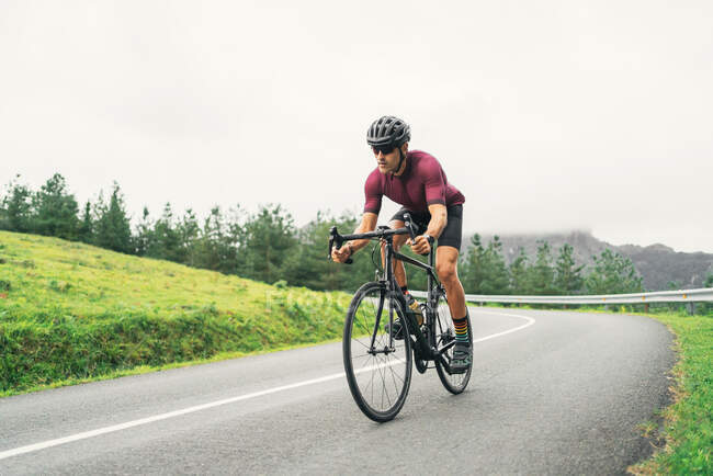 Sportivo in casco protettivo in bicicletta durante l'allenamento su strada asfaltata contro la collina verde e gli alberi sotto il cielo chiaro — Foto stock