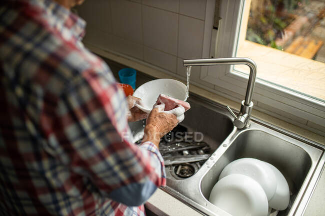 Вид сбоку обрезанного неузнаваемого мужчины, моющего грязные пластинки, стоя рядом с раковиной на кухне и выполняя домашнюю работу — стоковое фото