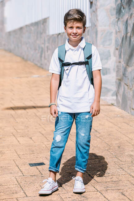 Содержание школьника в разорванных джинсах и резиновых туфлях, смотрящего в камеру на мощеной тротуаре в солнечном городе — стоковое фото