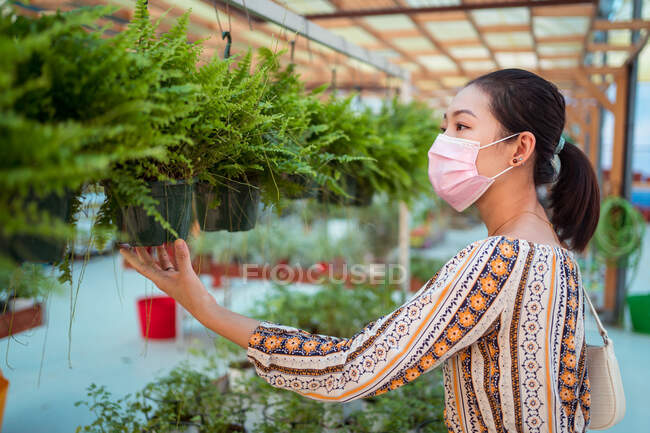Vista laterale di giovane acquirente femminile etnico in maschera sterile scegliendo pianta in vaso nel centro del giardino — Foto stock