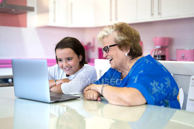 Nieta sonriente y abuela sentadas en la mesa y viendo videos en la computadora portátil en la cocina ligera en el apartamento - foto de stock