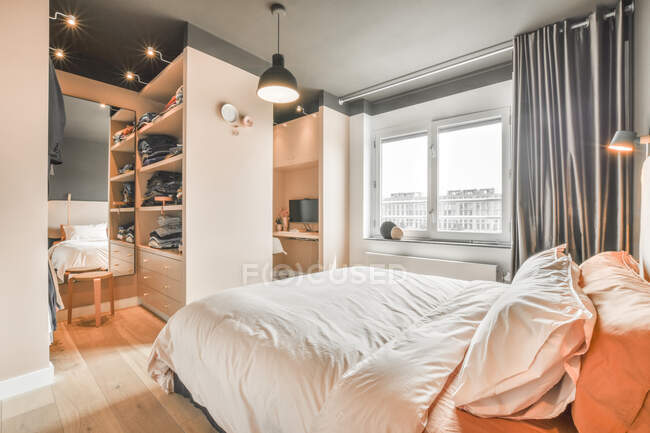 Удобная кровать с легким бельем, расположенная напротив шкафа и зеркала в стильной спальне в дневное время — стоковое фото