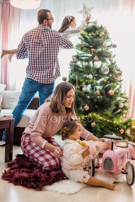 Aufgeregter Vater mit Tochter auf Händen schmückt Tanne mit Stern im Wohnzimmer mit Mutter und Kleinkind beim Spielen mit Spielzeug und Velomobil — Stockfoto