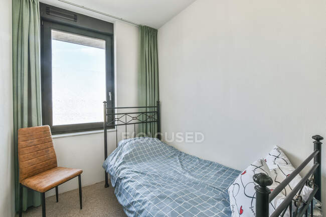 Interior do quarto pequeno com cama sob cobertor e travesseiros perto de cadeira e janela em plano — Fotografia de Stock