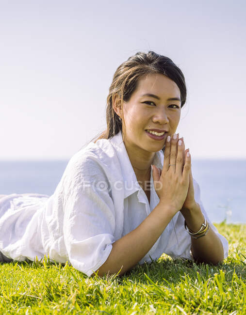 Aufrichtige junge ethnische Frau in Freizeitkleidung blickt in die Kamera, während sie tagsüber an der grünen Küste liegt — Stockfoto
