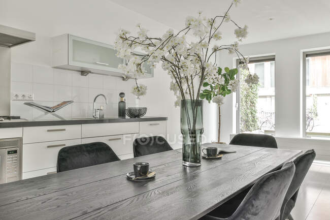 Interno della cucina luminosa contemporanea e zona pranzo con grande tavolo con mazzo di fiori e sedie in appartamento moderno durante il giorno — Foto stock
