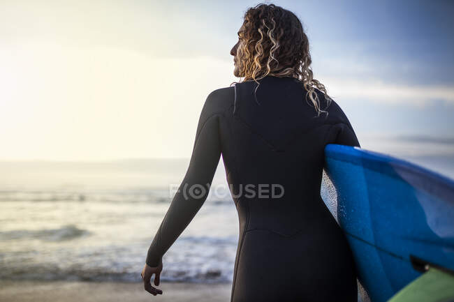 Vue arrière d'une jeune femme méconnaissable debout sur la rive avec une planche de surf avant d'entrer dans la mer au coucher du soleil sur la plage des Asturies, Espagne — Photo de stock