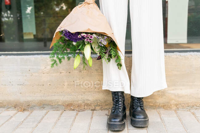 Cultivez une femelle fraîche méconnaissable dans un pantalon et des bottes en cuir debout avec un bouquet floral fleuri dans un emballage sur un trottoir carrelé — Photo de stock