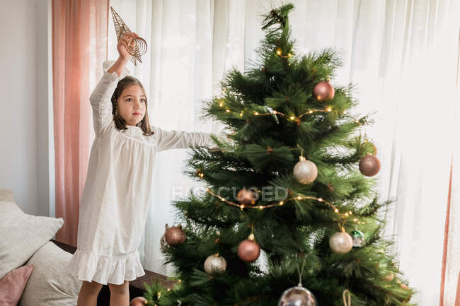Серйозна дівчина стоїть на дивані і прикрашає хвойну ялинку зіркою для святкування свята вдома — стокове фото