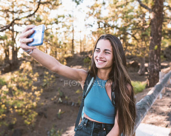 Cheerful cool female teenager with rucksack taking selfie while sitting on fence in Tenerife Spain — Stock Photo