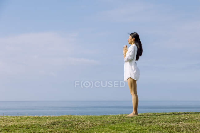 Side view of mindful young ethnic female with namaste hands practicing yoga on green coast against ocean in sunlight — Stock Photo