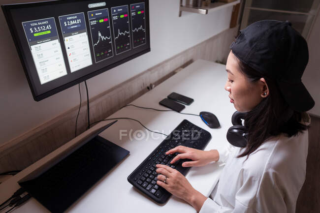 Visão lateral de alto ângulo da mulher asiática concentrada trabalhando no computador com gráficos mostrando dinâmica de mudanças no valor da criptomoeda no local de trabalho conveniente — Fotografia de Stock