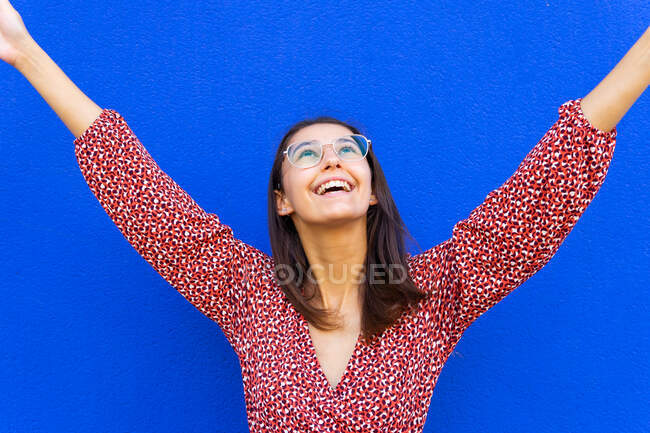 Giovane donna positiva con i capelli castani in abito rosso in piedi con le braccia alzate e guardando sullo sfondo blu durante il giorno — Foto stock