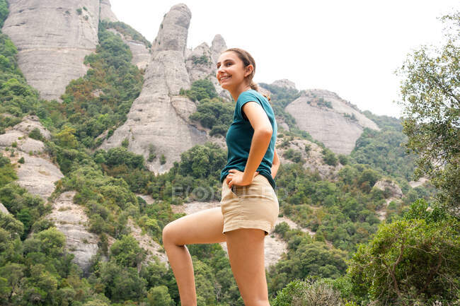 Vue latérale d'une voyageuse joyeuse avec les mains sur les hanches contemplant Montserrat avec des arbres tout en regardant loin lors d'une excursion en Espagne — Photo de stock