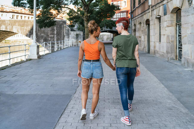 Vista posterior de mujeres homosexuales jóvenes con tatuajes tomados de la mano mientras caminan por la pasarela en la ciudad - foto de stock