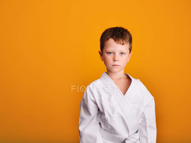 Ernsthafter Junge im weißen Kimono zum Üben eines Karate-Angriffs steht vor leuchtend orangefarbenem Hintergrund und blickt in die Kamera — Stockfoto
