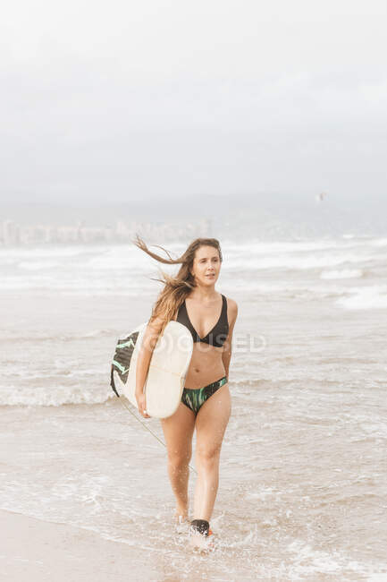 Giovane atleta donna in costume da bagno con capelli volanti e tavola da surf che cammina e distoglie lo sguardo sulla costa dell'oceano — Foto stock