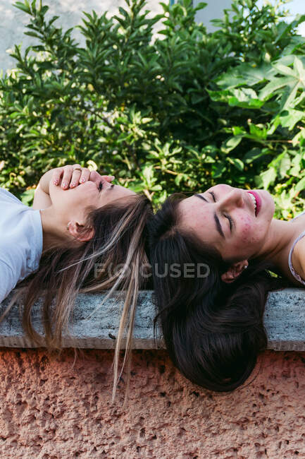 Vista lateral de una adolescente sonriente con acné acostada contra la mejor amiga con los ojos cerrados en el banco en el parque - foto de stock