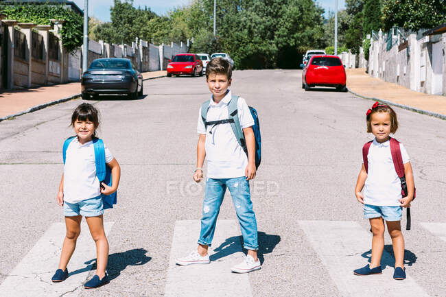 Schoolkids with rucksacks standing on asphalt road while looking at camera in sunny town — Stock Photo