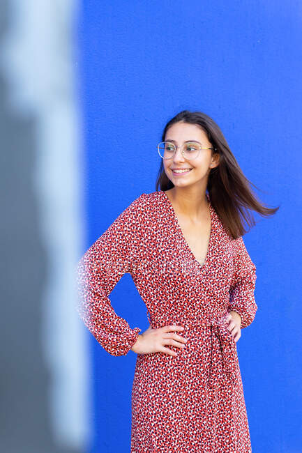 Happy female in stylish red dress standing with hands on waist against blue wall in daytime looking away — Stock Photo