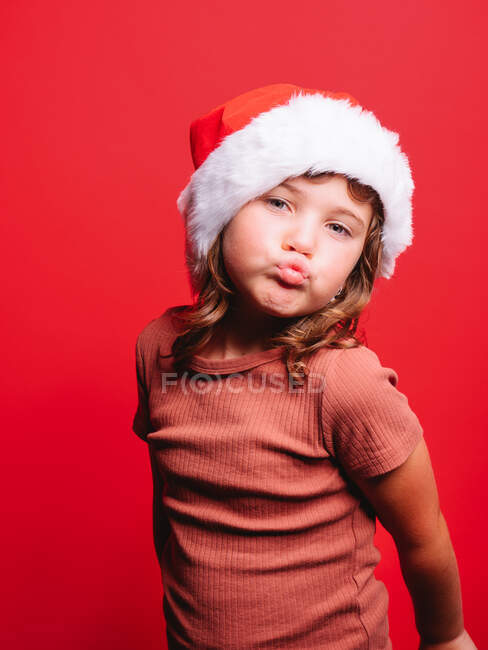 Adorable little girl in casual clothes and Santa hat pouting lips while standing against red background and looking at camera — Stock Photo