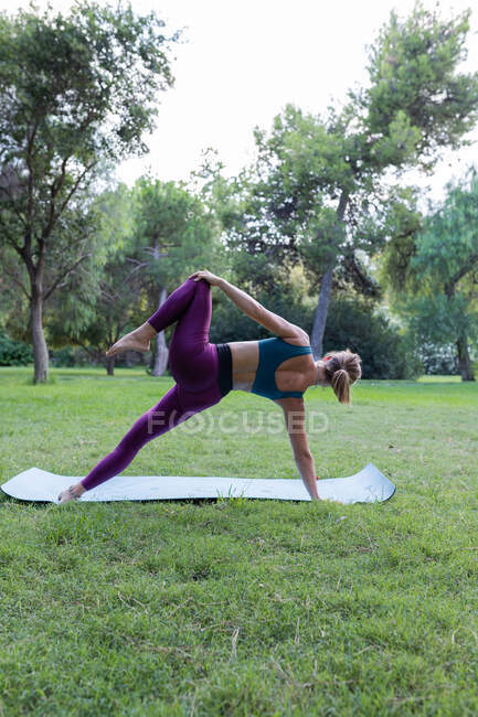 Вид ззаду на невизначену жінку У активному одязі роблять бічну дошку з піднятим коліном на килимку в зеленому парку в денний час — стокове фото