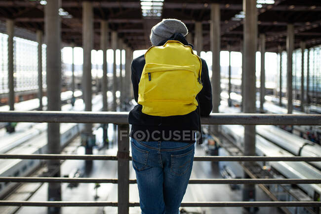 Rückansicht eines nicht wiederzuerkennenden männlichen Reisenden mit Rucksack, der am Geländer eines Durchgangs über Zügen am Bahnhof steht — Stockfoto